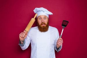 confused chef with beard and red apron chef holds wooden rolling pin photo