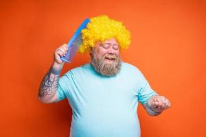 Fat happy man with beard, tattoos and sunglasses combs himself with a giant comb photo