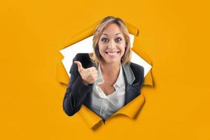 Happy caucasian girl with thumb up comes out of a hole on a yellow paper backdrop photo