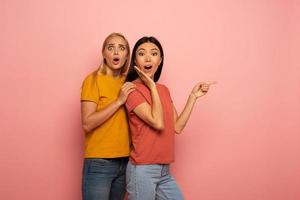 Two girls indicates something. Amazed expression face. Pink background with blank space for your text photo
