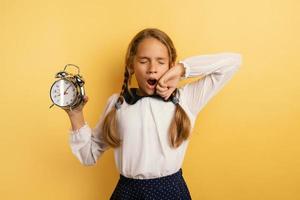 Young child student with ringing alarm clock is lazy going to school. Yellow background photo