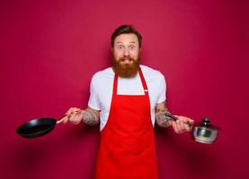 afraid chef with beard and red apron cooks with pan and pot photo