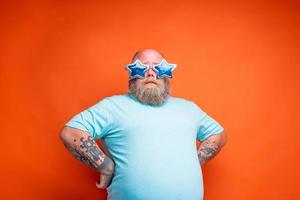 grasa pensativo hombre con barba, tatuajes y Gafas de sol es inseguro para alguna cosa foto