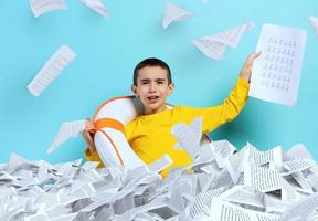 Young boy student is drowning in a sea of sheets of exercises. Cyan background photo