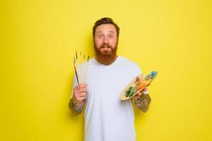 Undecided man with beard and tattoo is ready to draw with brushes photo