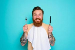 Happy man with tattoos is ready to eat with cutlery in hand photo