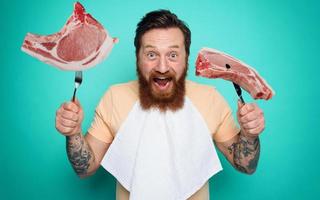 Man with tattoos is ready to eat meat with cutlery in hand. cyan background photo