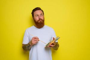 Undecided man with beard and tattoo is ready to draw with brushes photo