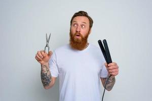 Undecided man with scissors and straightener is ready to work with hair photo