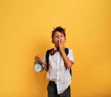 Young child student with ringing alarm clock is lazy going to school. Yellow background. photo