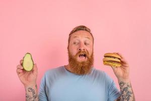 Hungry man with beard and tattoos is undecided if to eat an avocado or an hamburger photo