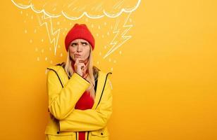 Girl thinks about negative things. Cloud, rain and lightning above her. Confused and pensive expression. Yellow background photo