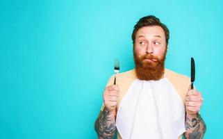 Man with tattoos is ready to eat with cutlery in hand. cyan background photo