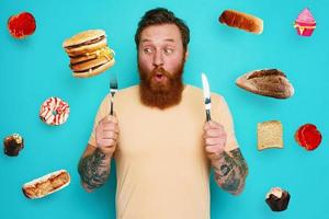 Man with tattoos is ready to eat sandwich and sweets with cutlery in hand. cyan background photo