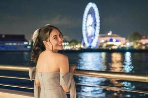 Beautiful young asian woman in dress smiling and sightseeing illuminated ferris wheel during cruise ship on the river in the night photo