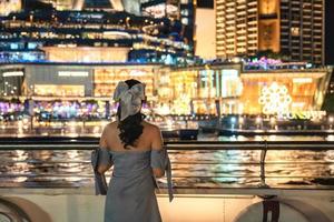 Beautiful young asian woman in dress looking at illuminated department store glowing in festival event on riverside in the night. Dinner cruise sailing and sightseeing on the Chao Phraya river photo