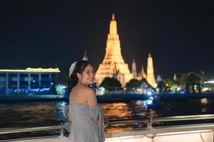 Beautiful young asian woman in dress standing and looking at camera on the deck with Wat Arun glowing in the night. Dinner cruise sailing on the Chao Phraya river photo