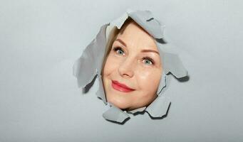 Surprised woman looking playfully in torn paper hole, has excited cheerful expression, looks through breakthrough of gray background. Wow, great news photo