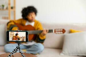 Blogger guitarist. African american girl blogger playing guitar talking to webcam recording vlog. Social media influencer woman streaming at home indoors. Music content creator broadcast tutorial. photo