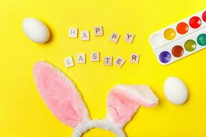 Inscription HAPPY EASTER letters eggs colorful paints and bunny ears isolated on trendy yellow background photo