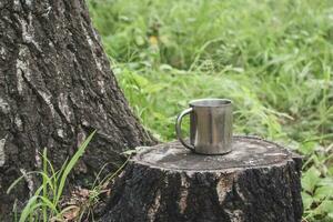 An iron tourist mug stands on a stump in a forest next to a thick tree against a background of blurred grass. Selective focus on the mug. The background is blurry. Place for text. photo