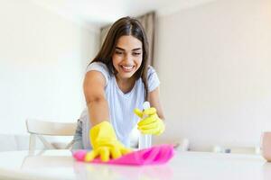 Beautiful young woman makes cleaning the house. Girl rubs dust. Smiling woman wearing rubber protective yellow gloves cleaning with rag and spray bottle detergent. Home, housekeeping concept. photo