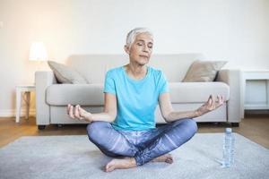 Senior woman practicing yoga, sitting in Half Lotus exercise, Ardha Padmasana pose, working out, wearing sportswear, meditation session, indoor full length, home interior photo