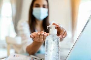 Young woman with face mask back at work in office after lockdown, disinfecting hands. Female employee in protective face mask sanitize hands with antibacterial liquid, protect from COVID-19 pandemic photo