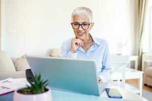 Focused old woman with white hair at home using laptop. Senior stylish entrepreneur wearing eyeglasses working on computer at home. Woman analyzing and managing domestic bills and home finance photo