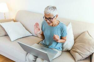 mujer madura feliz saludando a alguien mientras tiene una videollamada en una laptop en casa. anciana de cabello gris ondeando la mano frente a la computadora portátil mientras realiza una videollamada con los miembros de su familia. foto