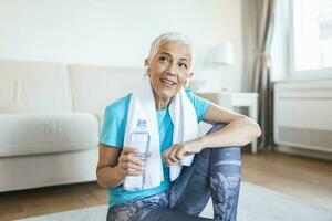 Elderly woman sitting on the mat, exhausted after the daily training. Senior woman taking a break while exercising at home. Athletic mature woman holding bottle of water photo