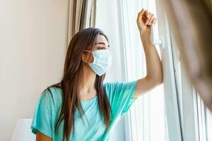 Sad lonely woman in isolation at home for virus outbreak or hypochondria standing next to the window during the day, looking through window. photo