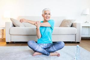 Senior woman doing warmup workout at home. Fitness woman doing stretch exercise stretching her arms - tricep and shoulders stretch . Elderly woman living an active lifestyle. photo