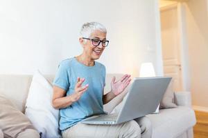 mujer madura feliz saludando a alguien mientras tiene una videollamada en una laptop en casa. anciana de cabello gris ondeando la mano frente a la computadora portátil mientras realiza una videollamada con los miembros de su familia. foto