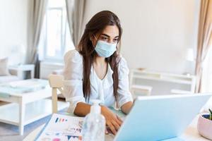 Quarantine, remote job and pandemic concept - woman wearing face protective medical mask for protection from virus disease with laptop computer having video call at home office photo