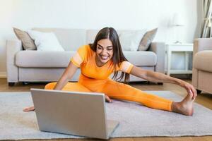 Girl training at home, doing workout and watching videos on laptop, training in living room. Beautiful woman is streching her legs. She is watching video on the internet and repeating the tasks photo