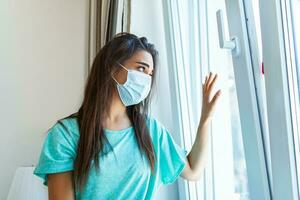 joven mujer vistiendo protector cara máscara mirando fuera de ventana con tristeza en su ojos, yo aislamiento debido a el global covid-19 coronavirus pandemia foto