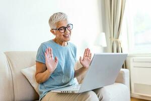 mujer madura feliz saludando a alguien mientras tiene una videollamada en una laptop en casa. anciana de cabello gris ondeando la mano frente a la computadora portátil mientras realiza una videollamada con los miembros de su familia. foto