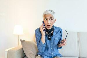 anciano hembra teniendo teléfono conversacion con su antiguo amigo utilizando Teléfono móvil, que se discute último Noticias con sorprendido rostro, sentado a hogar foto