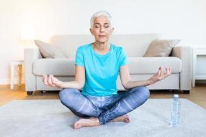 Senior woman practicing yoga, sitting in Half Lotus exercise, Ardha Padmasana pose, working out, wearing sportswear, meditation session, indoor full length, home interior photo