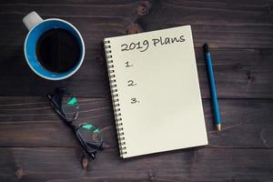 Notebook with 2019 plans massage, pencil, glasses and cup of coffee on wood background. photo