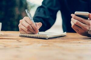 Man hand writing notebook paper and using phone on wood table in coffee shop with vintage toned filter. photo