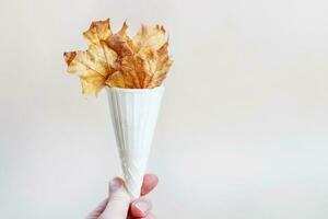 woman holding in hand autumn ice cream cone filled with autumn leaves on beige background. photo