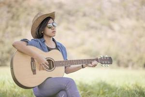 Women short hair wear hat and sunglasses sit playing guitar in grass field photo