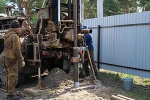 Team of workers with drilling rig on car are drilling artesian well for water in ground. Insertion of metal casing pipe into ground, installation of individual drinking supply, June 28, 2022, Russia, photo