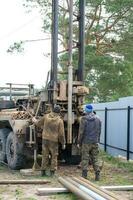 Team of workers with drilling rig on car are drilling artesian well for water in ground. Insertion of metal casing pipe into ground, installation of individual drinking supply, June 28, 2022, Russia, photo