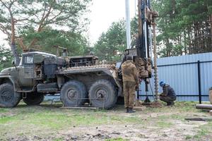 Team of workers with drilling rig on car are drilling artesian well for water in ground. Insertion of metal casing pipe into ground, installation of individual drinking supply, June 28, 2022, Russia, photo