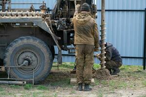 Team of workers with drilling rig on car are drilling artesian well for water in ground. Insertion of metal casing pipe into ground, installation of individual drinking supply, June 28, 2022, Russia, photo