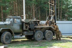 Team of workers with drilling rig on car are drilling artesian well for water in ground. Insertion of metal casing pipe into ground, installation of individual drinking supply, June 28, 2022, Russia, photo