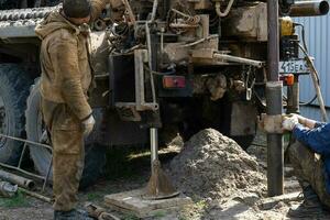 Team of workers with drilling rig on car are drilling artesian well for water in ground. Insertion of metal casing pipe into ground, installation of individual drinking supply, June 28, 2022, Russia, photo